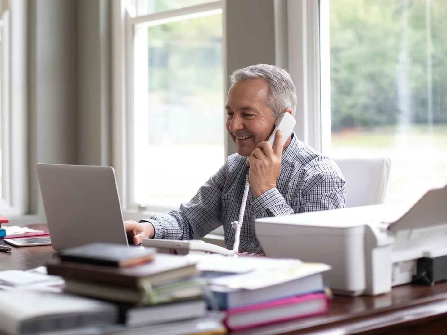 A smiling older man talking on a landline phone while working at his home office desk with a laptop.